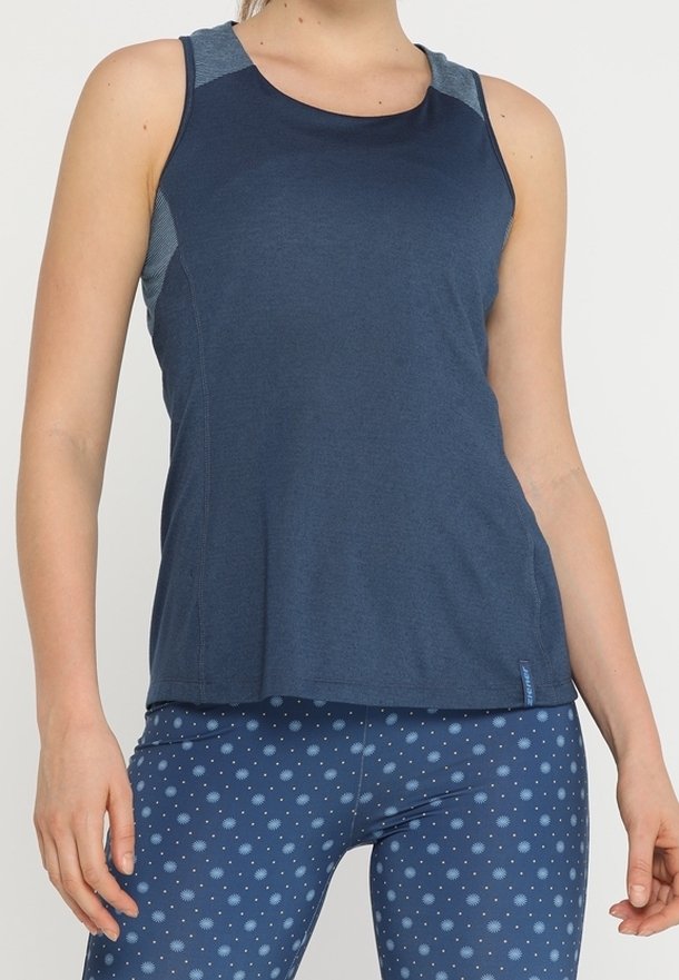 Femme portant un débardeur de sport bleu marine et un legging bleu orné d'un petit motif circulaire, debout bras détendus.
