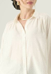 Biele button-up tričko so štruktúrovanou tkaninou, s klasickým golierom a voľným strihom. Viditeľné sú zlaté vrstvené náhrdelníky a okrúhle náušnice.