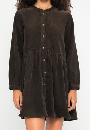 Frau trägt ein dunkelbraunes Cordkleid mit langen Ärmeln, Knopfleiste, geraffter Taille und rundem Ausschnitt.