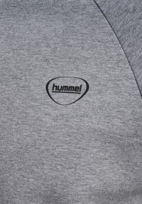 Grauer Stoff mit einem schwarzen "hummel"-Logo in einer unregelmäßigen abgerundeten Form, nahe einer Naht auf der oberen rechten Seite positioniert.