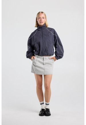 Blonde vrouw staat met handen in de zakken, draagt een donker jasje, lichtgrijze rok, witte sokken met zwarte strepen en zwarte schoenen.