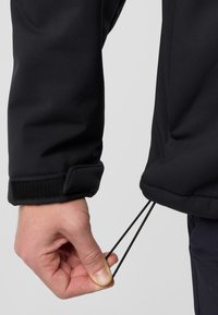 Veste noire au toucher lisse et à manches ajustées. Une main tire sur les cordons au bas pour l'ajuster. Fermeture à Velcro au niveau des poignets visible.