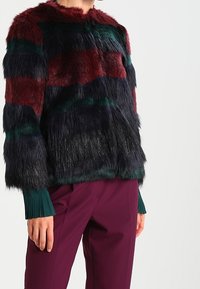 Faux fur jacket with horizontal stripes in burgundy, dark green, and black. Features a high collar and wide sleeves. Worn over purple pants.