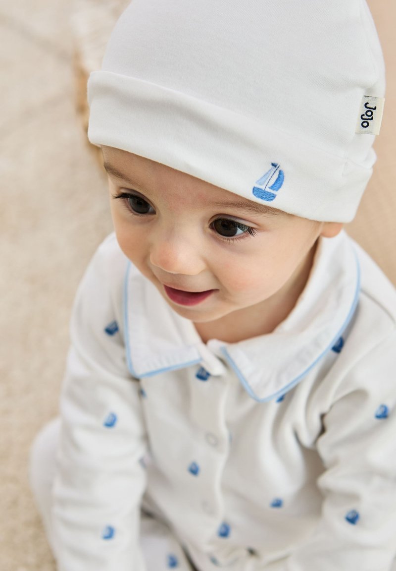 Witte babybeanie met een lichtblauwe zeilbootborduursel; de bijpassende witte pyjama's hebben kleine blauwe zeilbootpatronen en een blauwkleurig ontwerp.