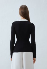 Pull en maille noire avec des manches longues, silhouette ajustée et détail de col blanc. Porté avec un pantalon large de couleur claire.