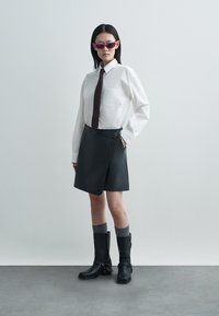 Λευκό πουκάμισο με κουμπιά, σκούρα φούστα τύπου wrap, γκρι κάλτσες και μαύρες μπότες μέχρι το γόνατο. Αξεσουάρ περιλαμβάνουν στενή σκουρόχρωμη γραβάτα και ροζ γυαλιά ηλίου.