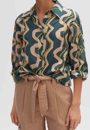 Knopfhemd mit blauen, beigen und gelben wellenförmigen Streifen, in hoch taillierte beige Hose mit einem vorne gebundenen Stoffgürtel gesteckt.