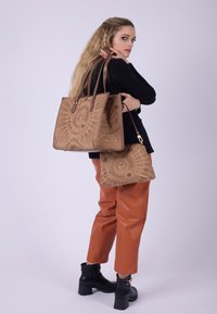Bruine tote bags met reliëfpatronen, een grote en een kleinere, gedragen door een model in oranje broek en zwarte enkellaarsjes, tegen een witte achtergrond.