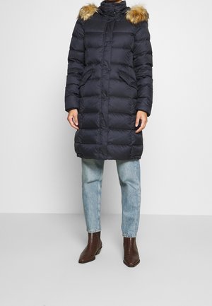 Personne portant un manteau matelassé noir longueur genoux avec capuche bordée de fourrure, jean bleu clair et bottines marron sur un fond uni.