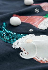 Schwarzer Stoff mit einem gestickten Gnom mit weißem Bart, einem türkisfarbenen Paillettenhut und weißen Bommel. Detaillierte Texturen und Muster sind vorhanden.