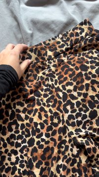 Leopárdmintás textil fekete, barna és krémszínű árnyalatokkal. Az anyag puhanak tűnik, rugalmas textúrával. Egy kéz pihen a derékszíjon.