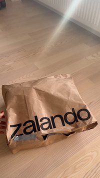 Hand med rött nagellack som håller ett ihopknycklat brunt Zalando-papperpaket på ett ljust trägolv nära en vit radiator.
