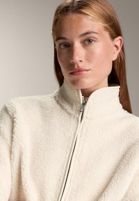 Giacca in pile crema con colletto alto e zip frontale. La texture è morbida e soffice, caratterizzata da un design semplice e minimal.