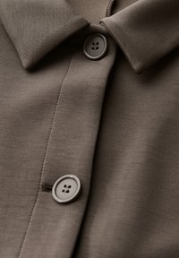 Brun skjorta med en klassisk krage, två grå knappar och en mjuk tygtextur med en subtil lyster.