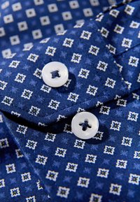 Donkerblauw overhemdstof met een geometrisch wit patroon, met twee witte knopen op een opgevouwen kraag waarop blauwe stikseldetails zichtbaar zijn.