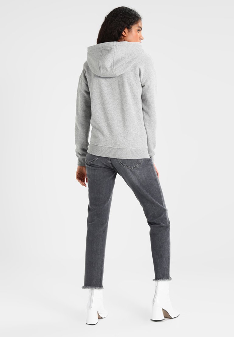 Femeie cu spatele, purtând un hanorac gri deschis, blugi gri închis cu tiv zdrențuit și cizme albe până la gleznă, pe un fundal simplu, deschis.