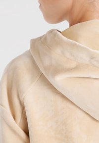Hoodie en velours beige avec une texture douce et une grande capuche. Présente un design d'épaule sans couture et un motif de tissu subtil.