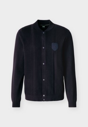 Juoda megzta striukė su mygtukais, su ribotu rankogaliu, apykakle, vertikaliomis tekstūrinėmis juostelėmis ir mažu siuvinėtu lipduku kairėje krūtinės pusėje.