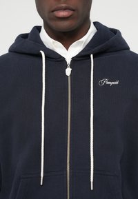 Detailný záber osoby v tmavomodrom zip up mikine s bielymi šnúrkami, strieborným zipsom a vyšitým nápisom "Pompeii" na hrudi.
