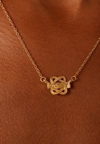 Collier en or présentant un pendentif en forme de nœud décoratif avec des détails intricats. Les maillons de la chaîne sont fins et délicats au toucher.