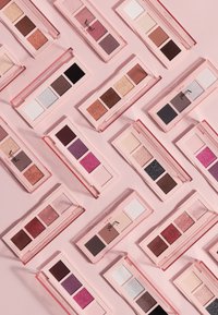 Verschiedene Lidschatten-Paletten in rosa Hüllen; mit glitzernden und matten Farbtönen in Braun, Pink, Weiß, Grau und Schwarz, auf einem hellrosa Hintergrund präsentiert.