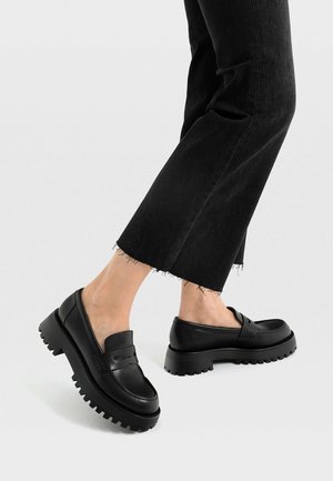 Ben iført korte sorte jeans og sorte læder loafers med chunky sål mod en ensartet lys baggrund.