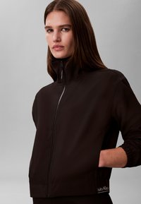 Sieviete ar gariem brūniem matiem, tērpusies melnā Calvin Klein rāvējslēdzēja džemperī ar augstu apkakli, rokas daļēji kabatās, fona krāsa neitrāla pelēka.