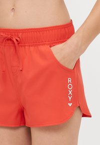 Rde kopalne hlače z vrvico za prilagajanje pasu, stranskimi žepi in belim logotipom "ROXY". Gladka tekstura in rahlo ukrivljen rob.