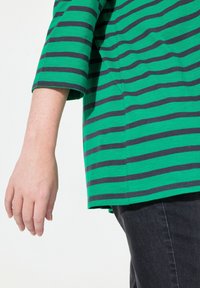 Grün gestreiftes Hemd mit dunkelblauen Horizontalstreifen; ausgestattet mit einem entspannten Schnitt, 3/4-Ärmeln und weichem Baumwollmaterial. Dunkle Jeans sind sichtbar.