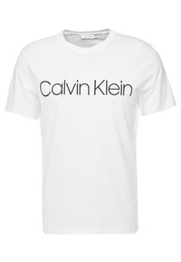 Bílý bavlněný tričko s kulatým výstřihem a krátkými rukávy, na kterém je výrazně zobrazeno "Calvin Klein" tučným černým písmem přes hruď.