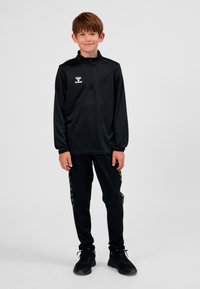 Junge steht und lächelt, trägt ein schwarzes Langarm-Zip-up-Oberteil und passende schwarze Hose mit schwarzen Turnschuhen vor einem schlichten Hintergrund.