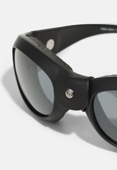 Gafas de sol negras con un acabado mate, que presentan lentes oscuros, detalles de cuero texturizado y un emblema metálico en la montura.