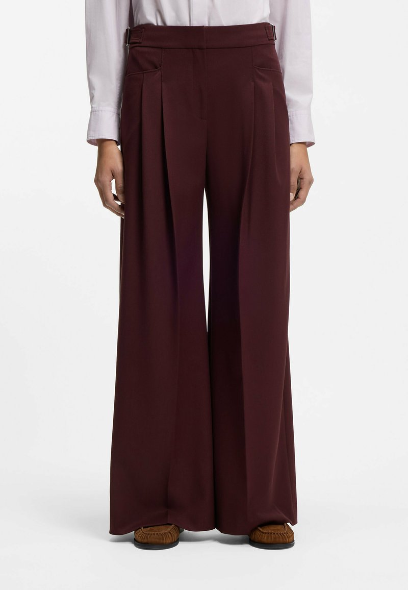 Personne portant un pantalon marron foncé taille haute, jambes larges, avec des plis, associé à une chemise blanche à manches longues rentrée dans le pantalon et des chaussures marron.