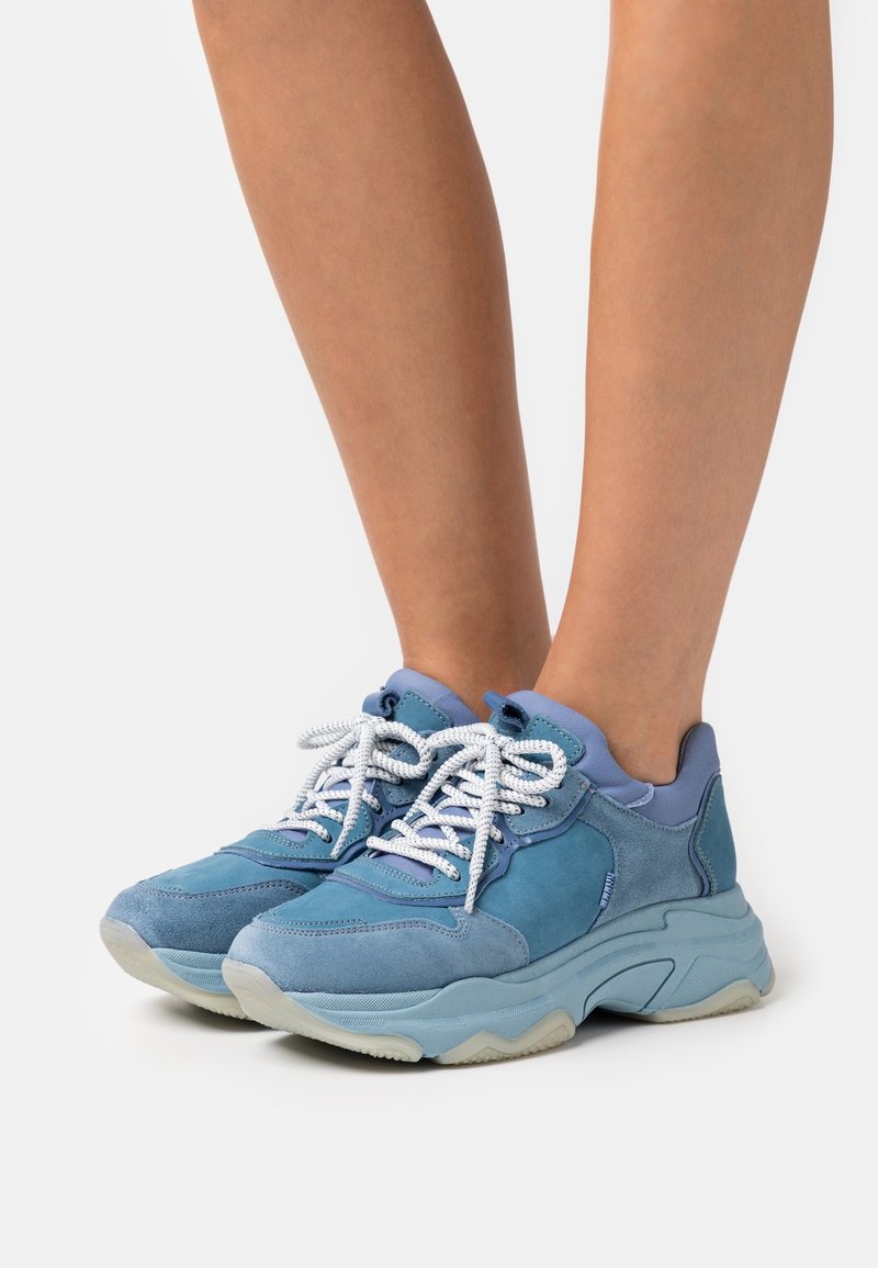 Modrá teniska s kombinací semišových a hladkých materiálů, vybavená bílými tkaničkami, robustní podrážkou a kontrastními světle modrými akcenty.