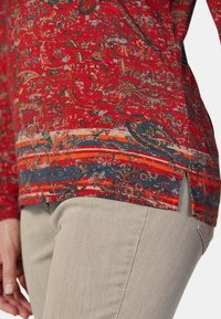 Rotes Oberteil mit floralem und abstraktem Muster, mit marineblauen und orangenen Streifen am Saum. Helle beigefarbene Hose mit sichtbarem Seitenschlitz.
