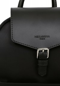Zaino nero con un ampio risvolto in pelle liscia, dotato di una fibbia argentata e dettagli cuciti. Logo "HEXAGONA PARIS" impresso sul risvolto.