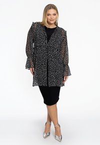 Transparante zwarte jacket met luipaardprint en ruches aan de mouwen, gedragen over een aansluitende zwarte jurk en gecombineerd met zilveren puntige hakken.