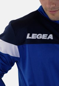 Blauwe sportjacket met een donkerblauwe bovenpaneel, witte accenten en geborduurd "LEGEA"-logo, vervaardigd uit glad, lichtgewicht materiaal.