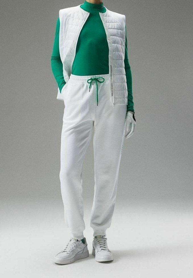 Personne portant un haut vert à manches longues, un gilet matelassé blanc, un pantalon de jogging blanc, des baskets blanches et un gant blanc à une main.