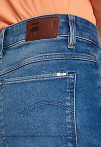 Jeans de mezclilla azul oscuro con un parche de cuero marrón con logo en relieve en la pretina y detalles sutiles de costura en los bolsillos.