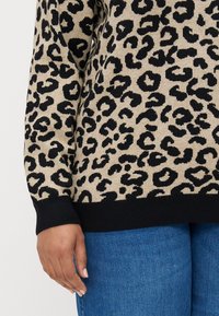 Leopardmønstret genser i beige med sorte flekker. Har en ribbestrikket svart nederkant og mansjetter. Kombinert med blå denim. Nærbilde av ermet og hånden.