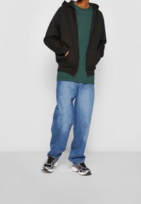 Sudadera negra con cremallera y bolsillos frontales, camiseta verde debajo, pantalones vaqueros holgados de color azul claro y zapatillas plateadas/negras. Look sencillo y casual.