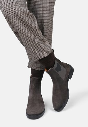 Person wearing brown checkered pants, dark brown socks, and dark gray suede Chelsea boots with a black heart detail on the elastic side.