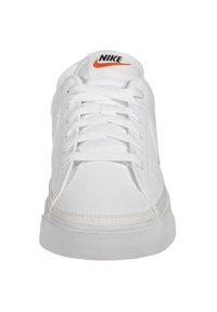 Bílé tenisky Nike s texturovaným koženým svrškem, s bílými šňůrkami, polstrovaným jazykem a černo-oranžovým logem na jazyku.