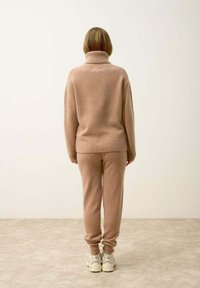 Beige neulepusero, jossa on korkeakaulus, yhdistettynä samansävyisiin kapeisiin housuihin. Molemmissa vaatekappaleissa on pehmeä rakenne ja rento istuvuus.