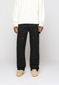 Black cargo pants with large pockets and snap fasteners, made from a sturdy material. Paired with a cream-colored long-sleeve shirt.
