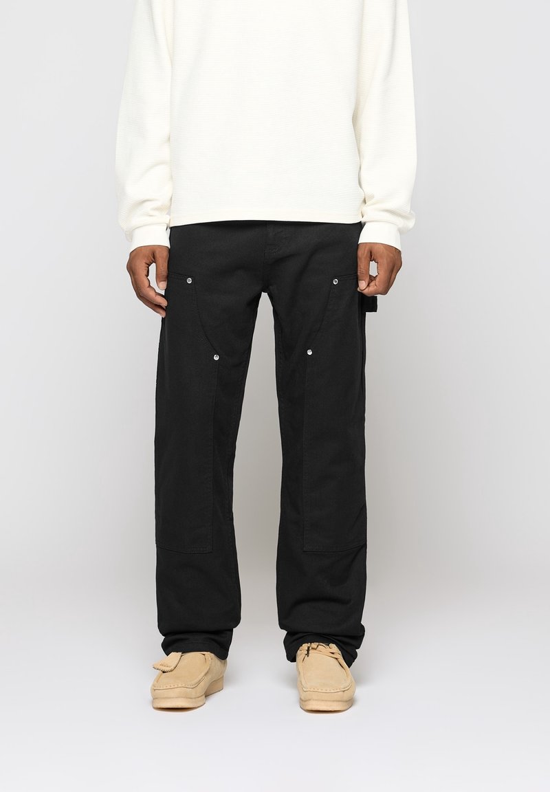 Black cargo pants with large pockets and snap fasteners, made from a sturdy material. Paired with a cream-colored long-sleeve shirt.