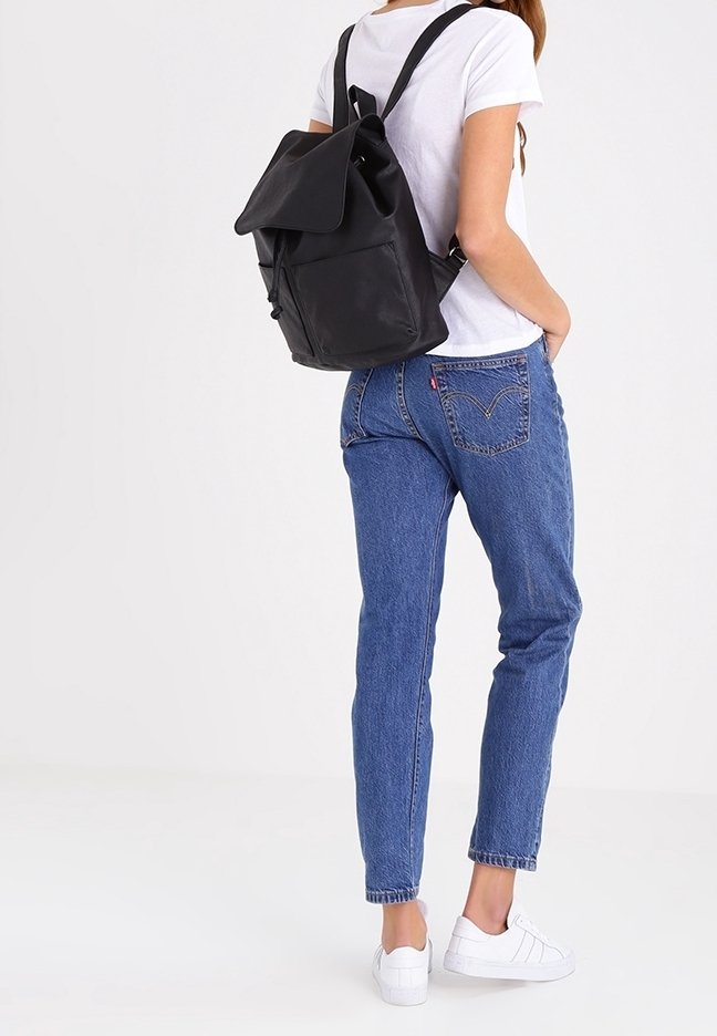 Sac à dos en cuir noir avec rabat pliant, bretelles réglables et poche avant. Porté avec un t-shirt blanc, un jean bleu et des baskets blanches.