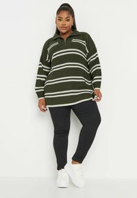 Stickad jumper i mörkgrön med vita ränder, med hög krage och dragkedja. Kombinerad med svarta byxor och vita sneakers.