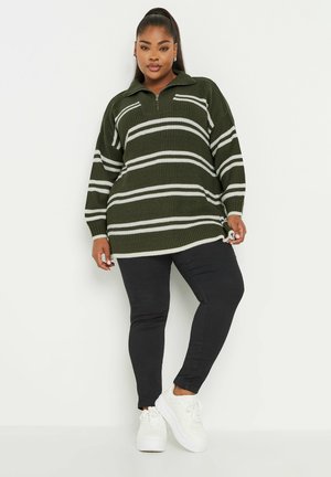 Suéter de punto en verde oscuro con franjas blancas, con cuello alto y cierre de cremallera. Combinado con pantalones negros y zapatillas blancas.